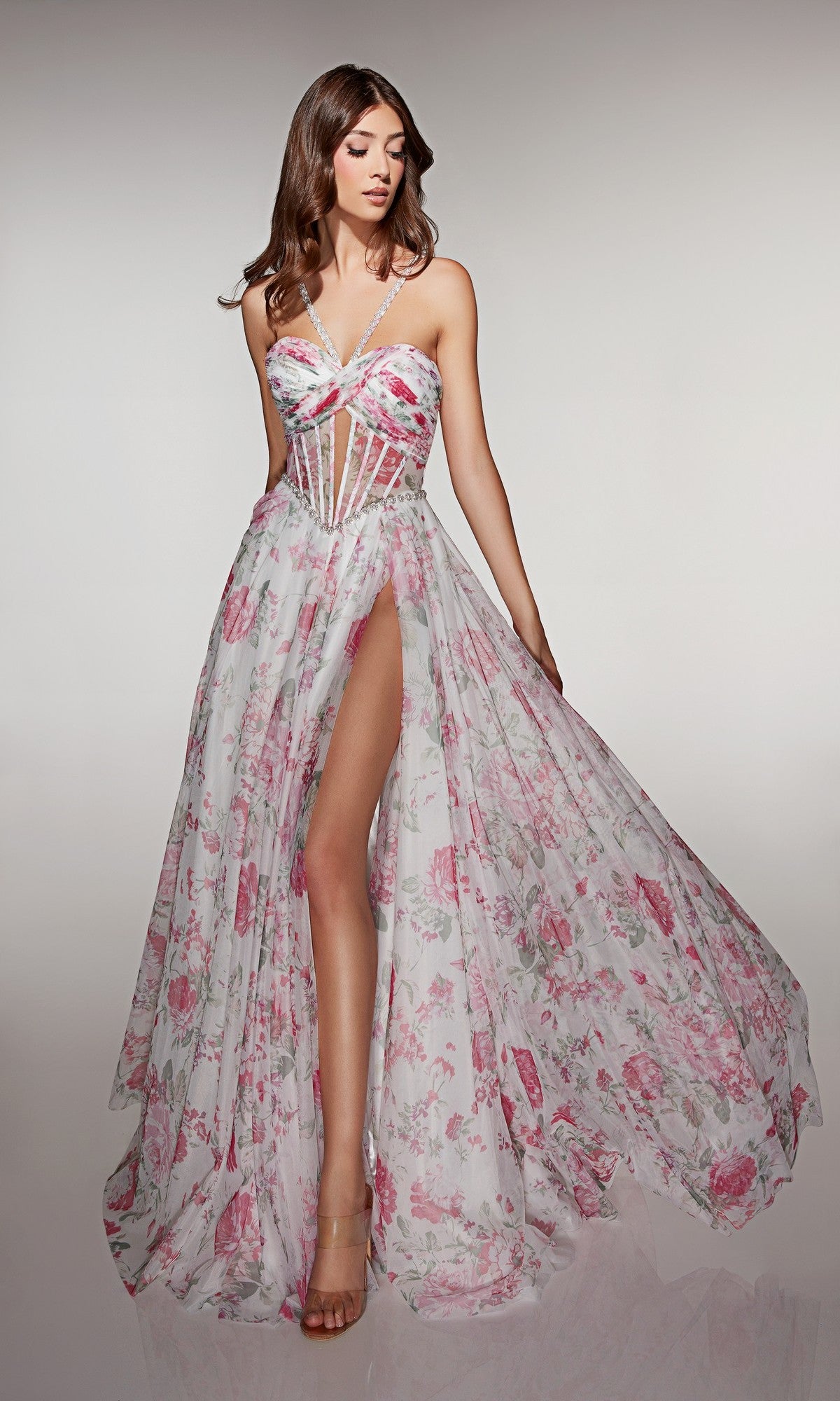 A woman wears the Alyce 61994 long prom dress—pink floral with a sheer corset bodice, spaghetti straps, and a high slit—standing against a neutral background.